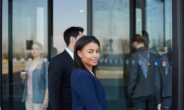 Female HR leader with short straight hair represents a PE-backed company CHRO as they walk through revolving glass door to enter office building with 4 colleagues wearing professional blazers.