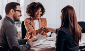 Female human resources executive leader CHRO 2 female shaking hands with with new hire across the desk wearing a suit jacket and smiling over papaerwork being signed. To the left is a male smiling at the human resources leader. Featured female has curly hair in the shape of a lose textured afro that cuts off at their chin length.